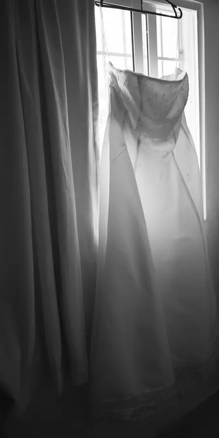 A wedding dress hanging in a tall window, soft morning light through the glass